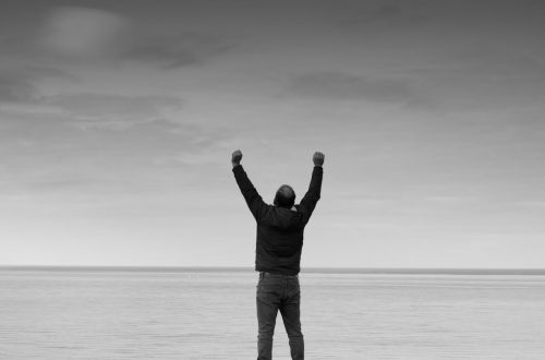 Black and white image of a person raising arms in celebration by the sea.