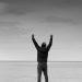 Black and white image of a person raising arms in celebration by the sea.