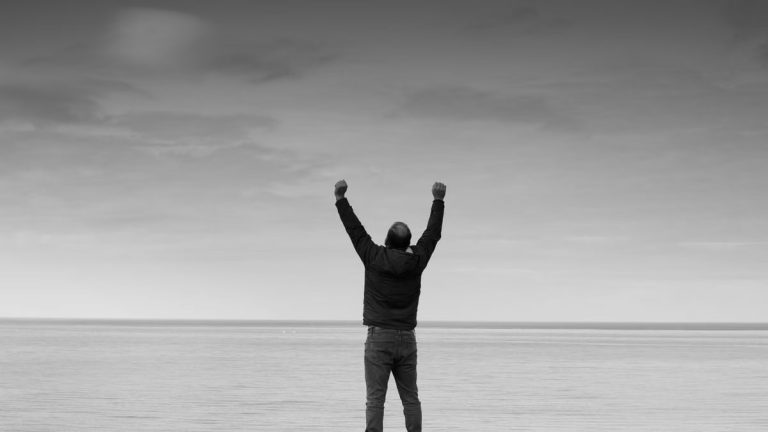 Black and white image of a person raising arms in celebration by the sea.